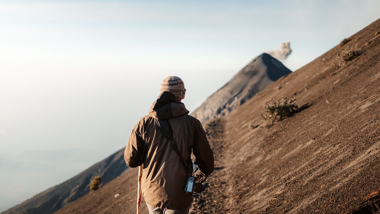 Hiker in winter coat walks toward erupting volcano on rocky terrain with misty sky backdrop.