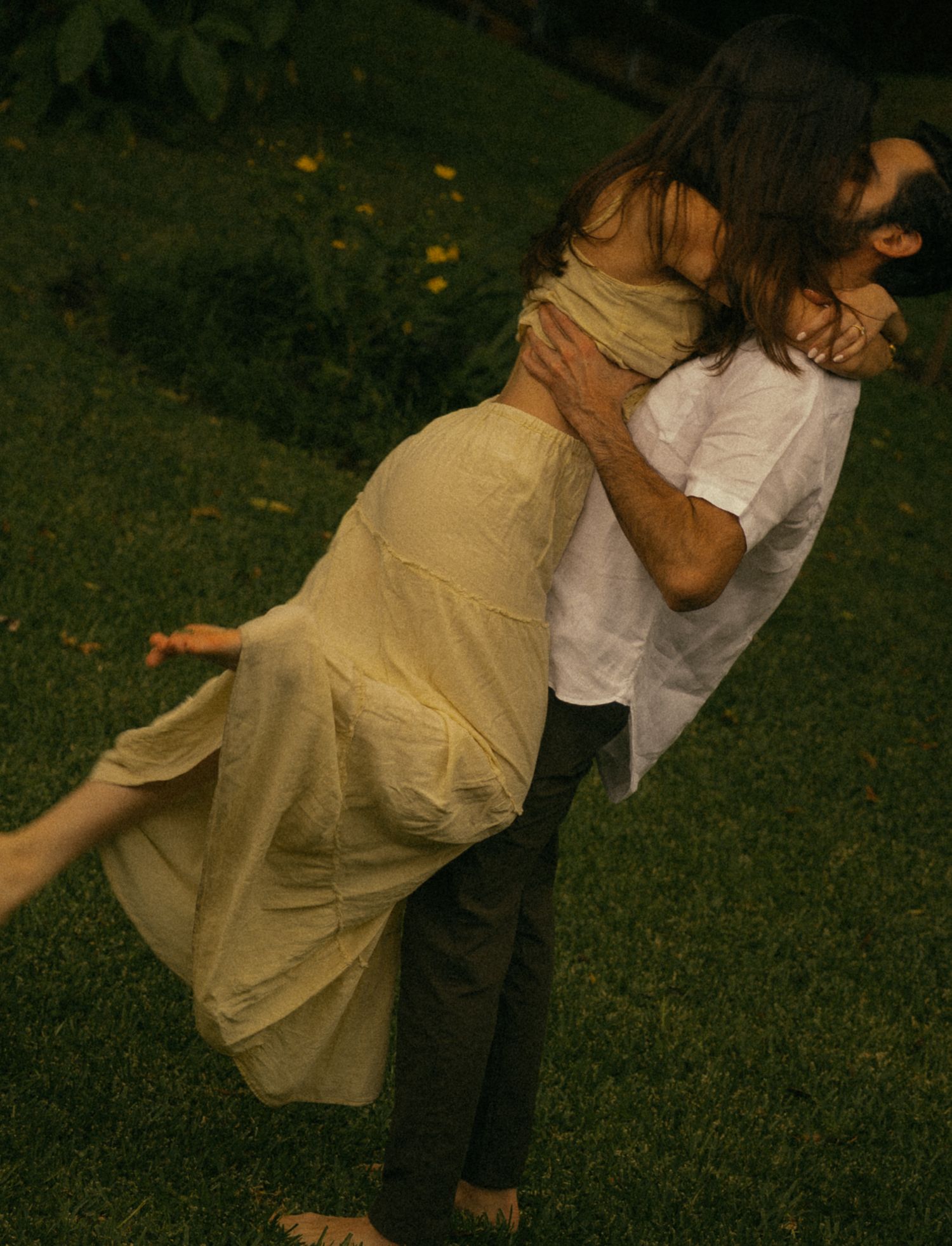 Couple embracing outdoors on green grass, man lifting woman in yellow dress.