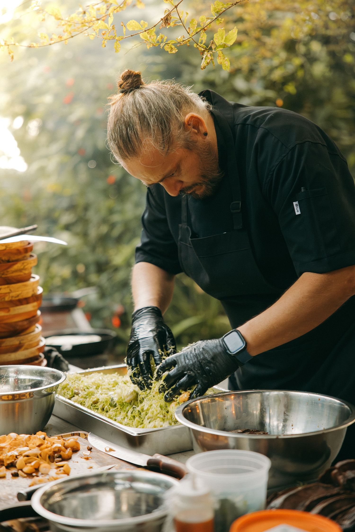 Chef in black uniform prepares fresh salad outdoors, wearing gloves at a rustic food station surrounded by bowls.