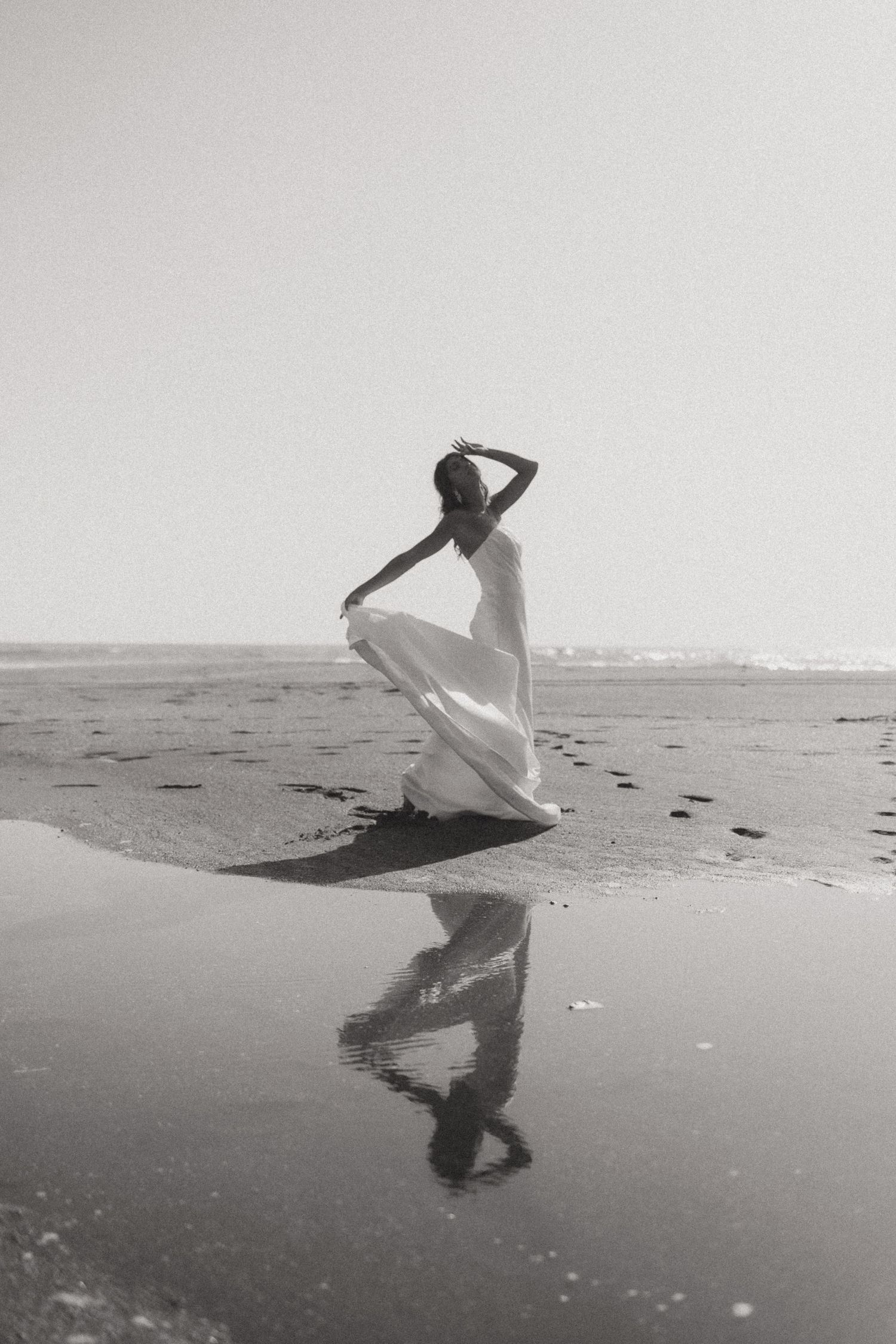 Black and white photo of a woman in a flowing white dress dancing on a reflective wet beach.