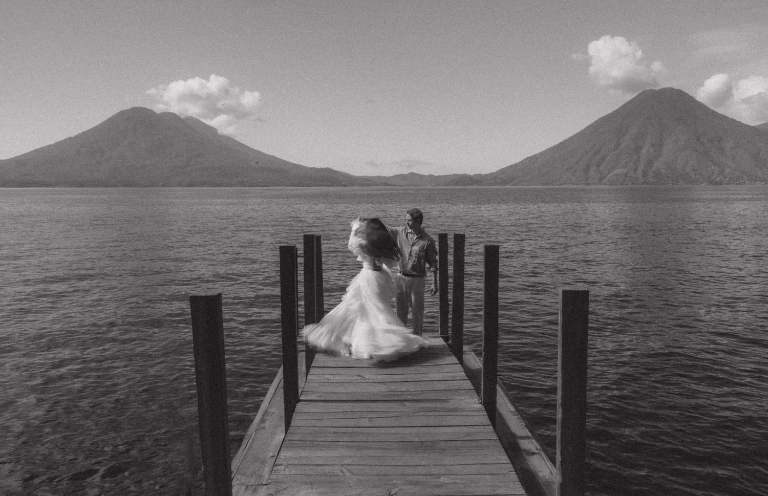 Couple in wedding attire standing on a wooden dock overlooking a serene lake with volcanic mountains in black and white.