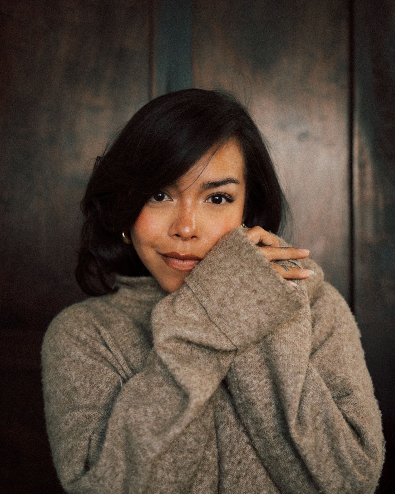 Young woman in cozy gray turtleneck sweater posing thoughtfully against a dark teal wooden background.