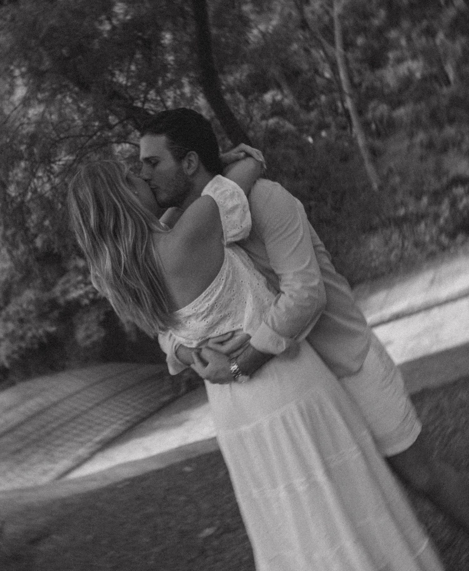 Black and white photo of a couple sharing a romantic kiss outdoors, woman in white dress embraced by man in light suit.