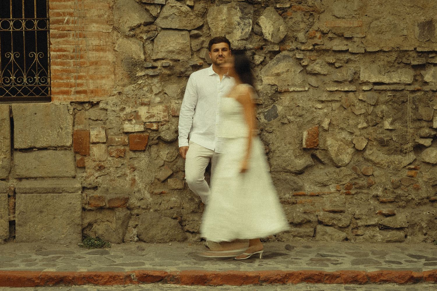 Couple in white outfits posing against a rustic stone wall, man in white shirt and pants, woman in elegant white dress.