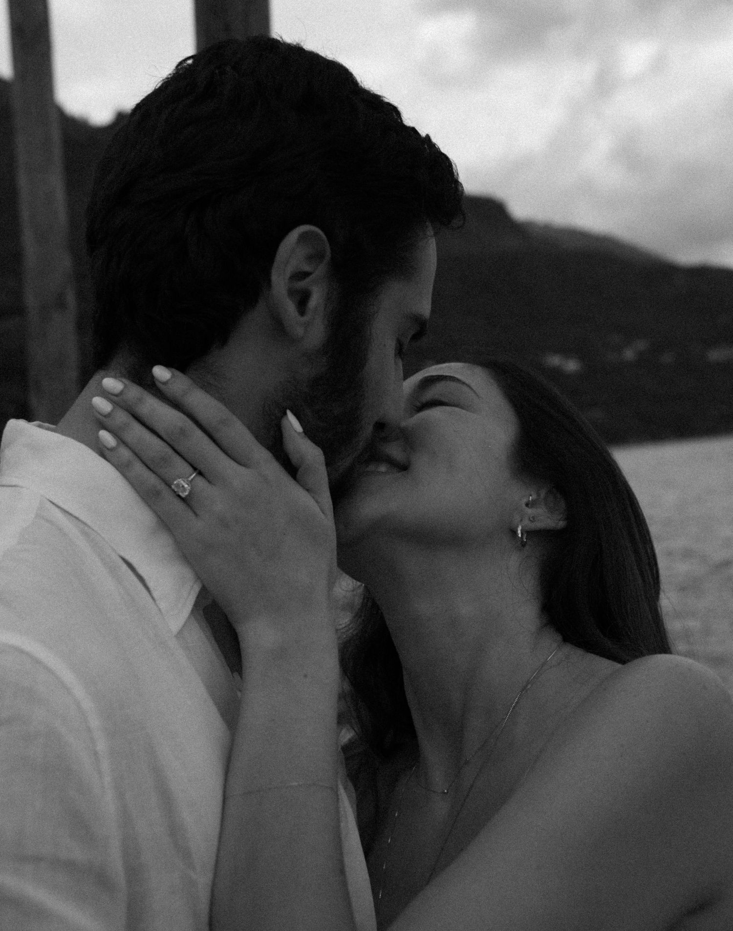 Black and white photo of a couple sharing a romantic kiss, woman wearing an engagement ring, outdoor waterfront setting.