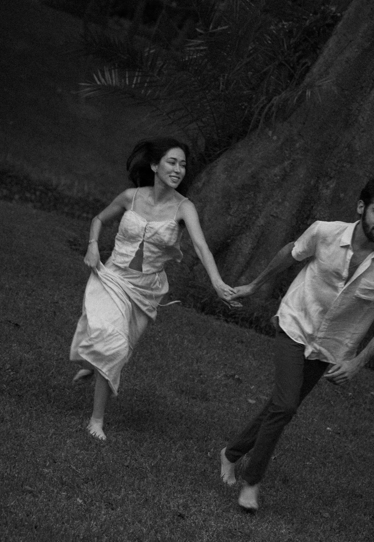A joyful couple runs barefoot through green grass near a large tree in a black and white romantic outdoor photo.