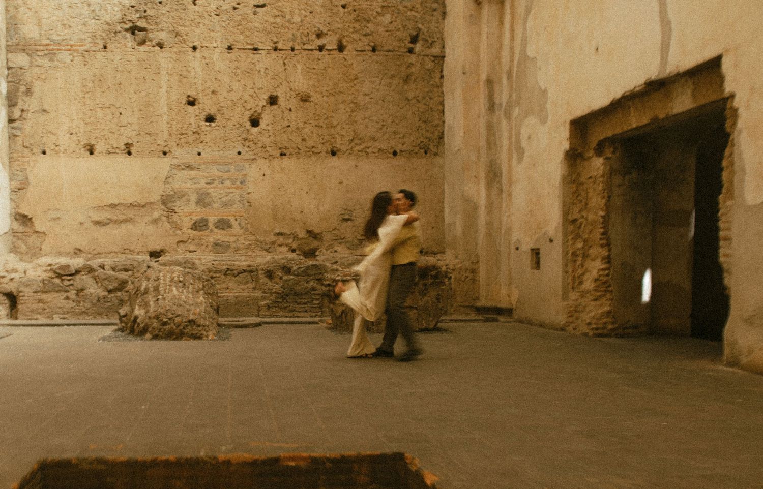 A couple embraces and dances in the center of a weathered, sunlit stone ruin with crumbling walls and an open doorway.