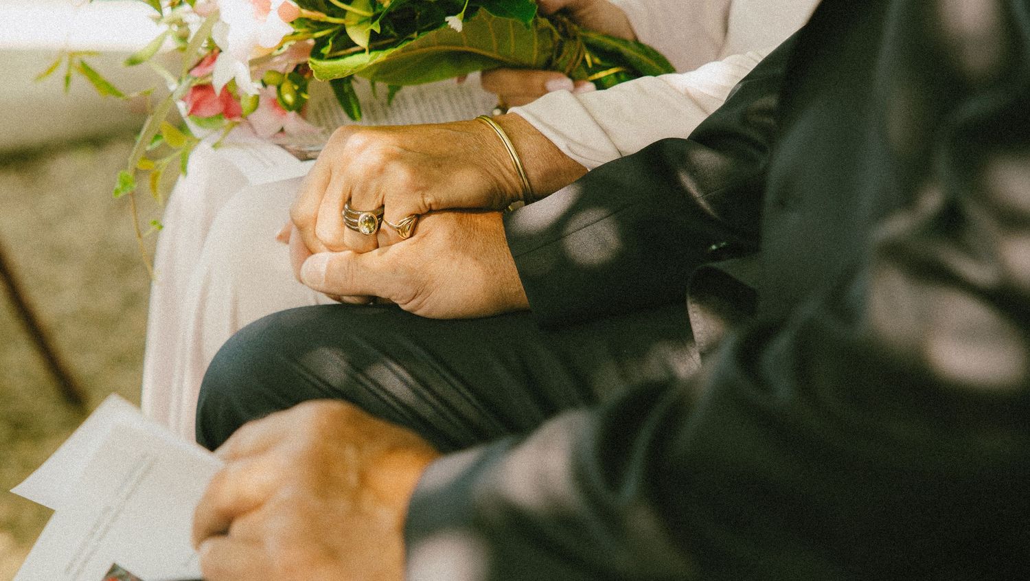 Elderly couple holding hands at wedding ceremony, bride holding pink bouquet, groom in black suit.