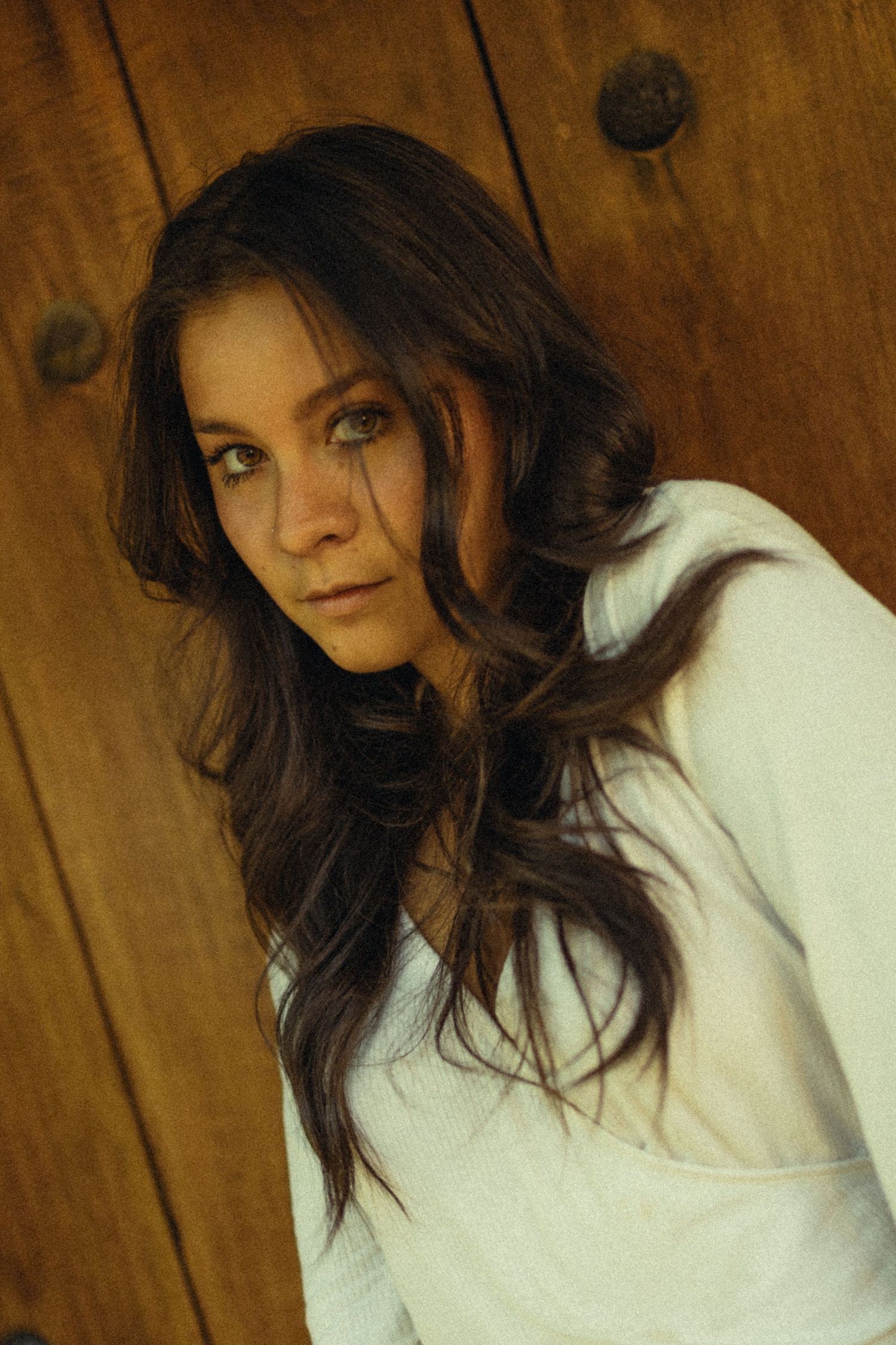 Young woman with long dark wavy hair in white top leaning against rustic wooden wall, looking over shoulder.