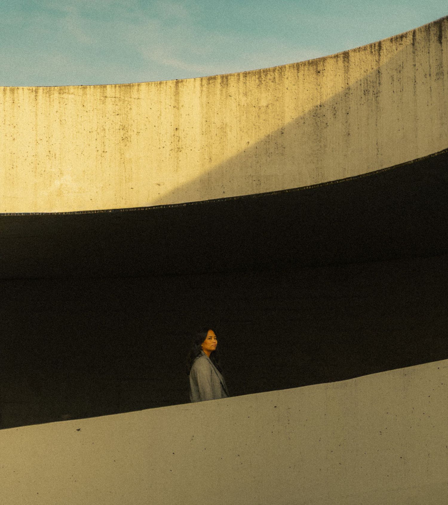 Person standing between curved concrete walls with dramatic shadow stripes and blue sky above.