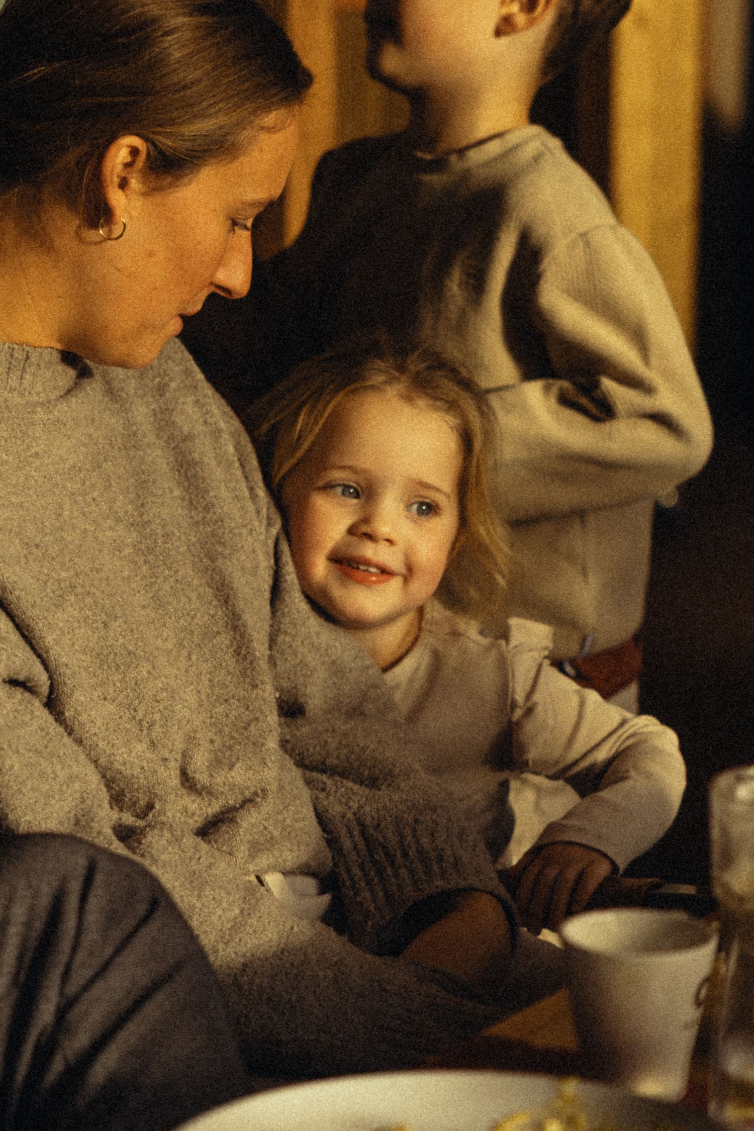 A woman in a gray sweater sits with a smiling blonde toddler girl, while another child stands in the background.