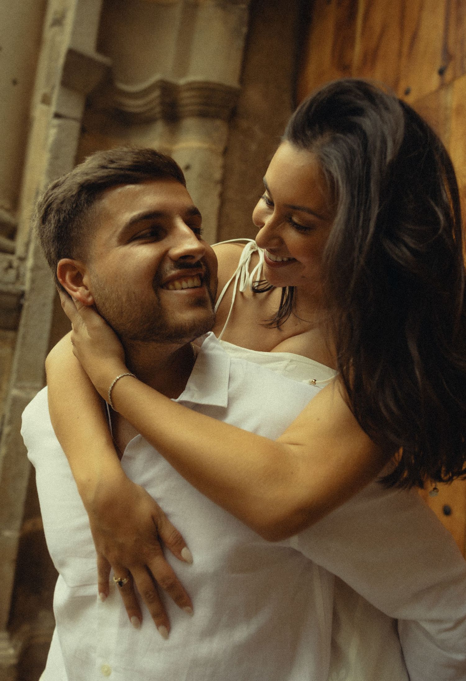 A smiling couple in white outfits embrace warmly in front of a rustic wooden door, sharing a joyful romantic moment.