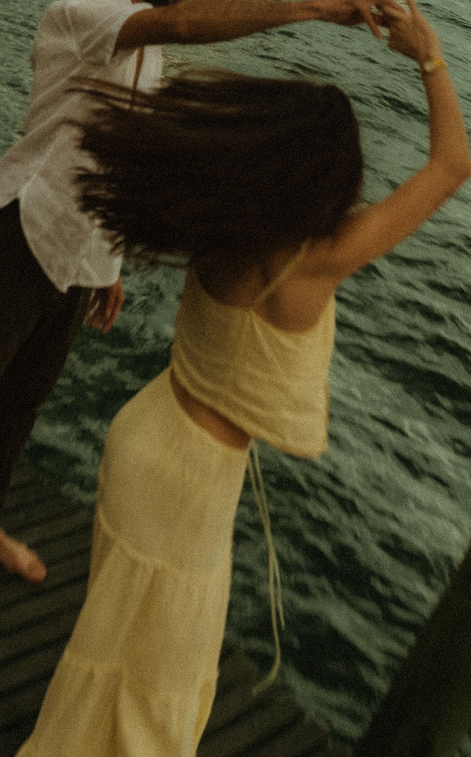 A woman in a yellow dress twirls gracefully near water, her dark hair flowing as a man in white holds her hand.
