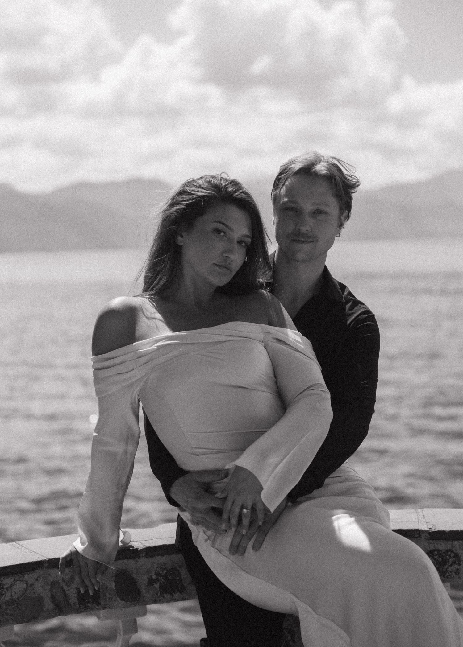 Couple poses by lakeside railing with mountain backdrop in elegant black and white engagement photo.