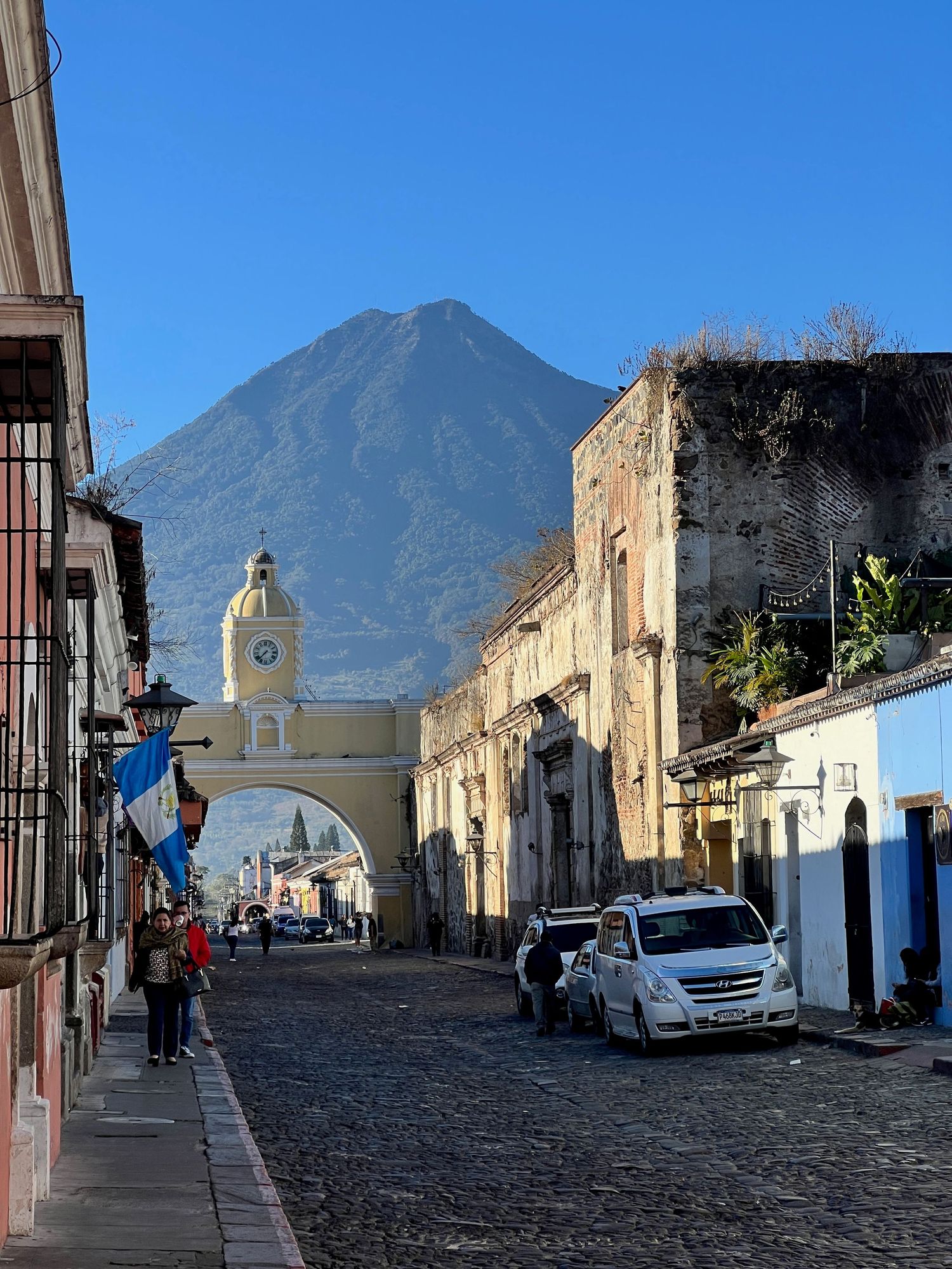 Cobblestone street in Antigua Guatemala with iconic yellow Santa Catalina Arch and Agua Volcano under clear blue sky.
