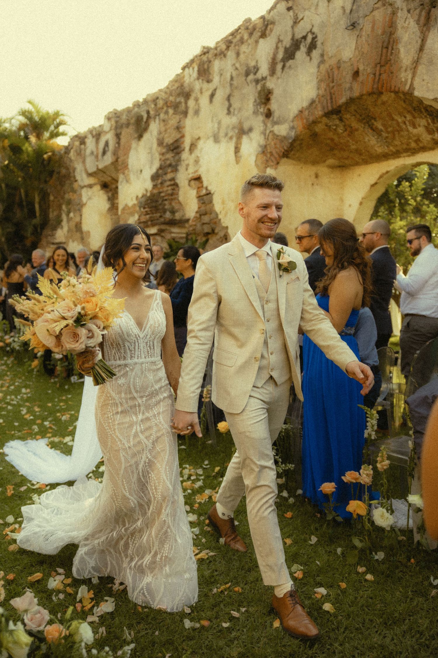 Smiling newlyweds walk down the aisle holding hands, bride in lace gown with yellow bouquet, groom in beige suit.