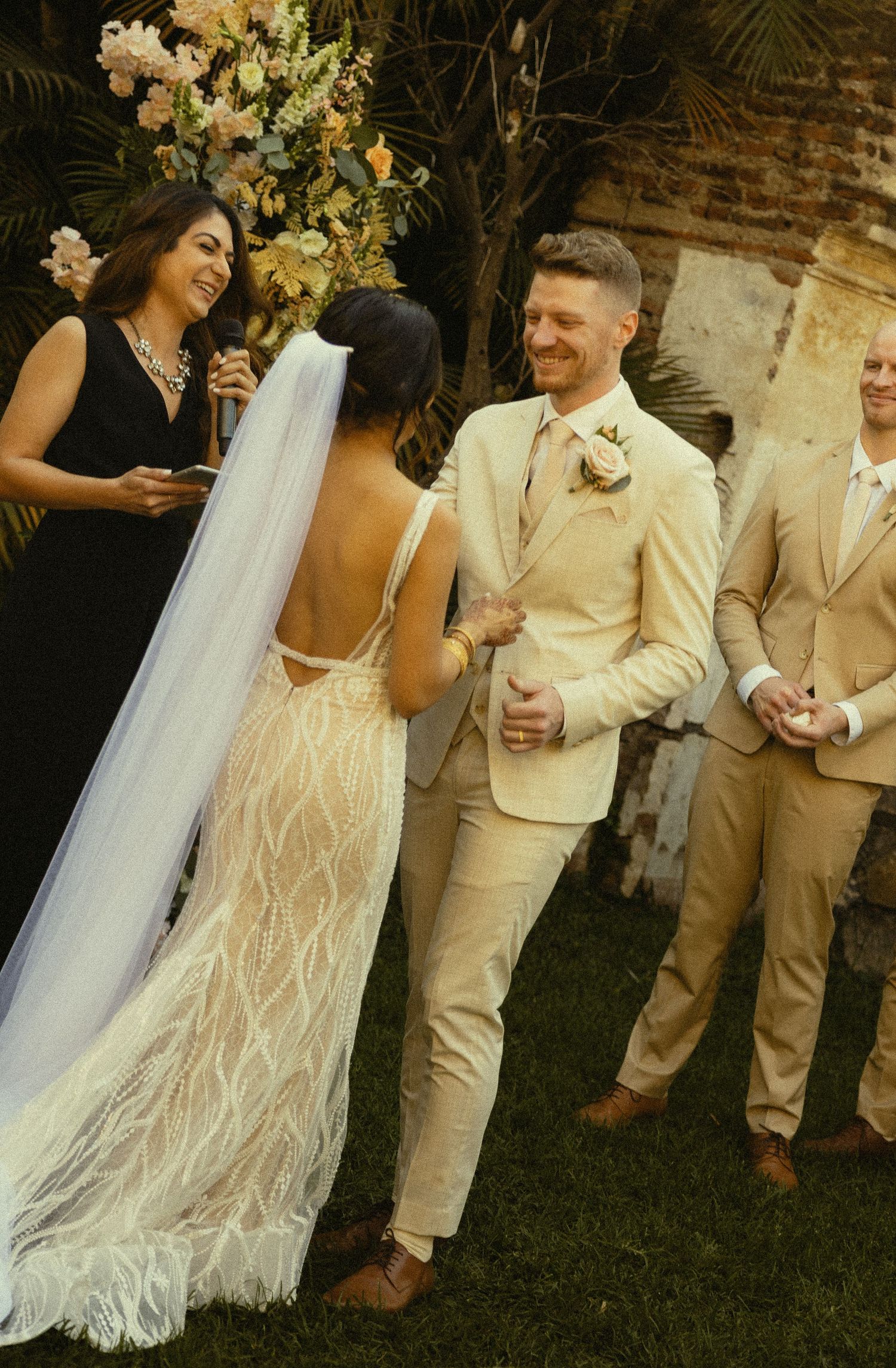Bride in backless lace gown and veil exchanges vows with groom in beige suit at outdoor wedding ceremony.