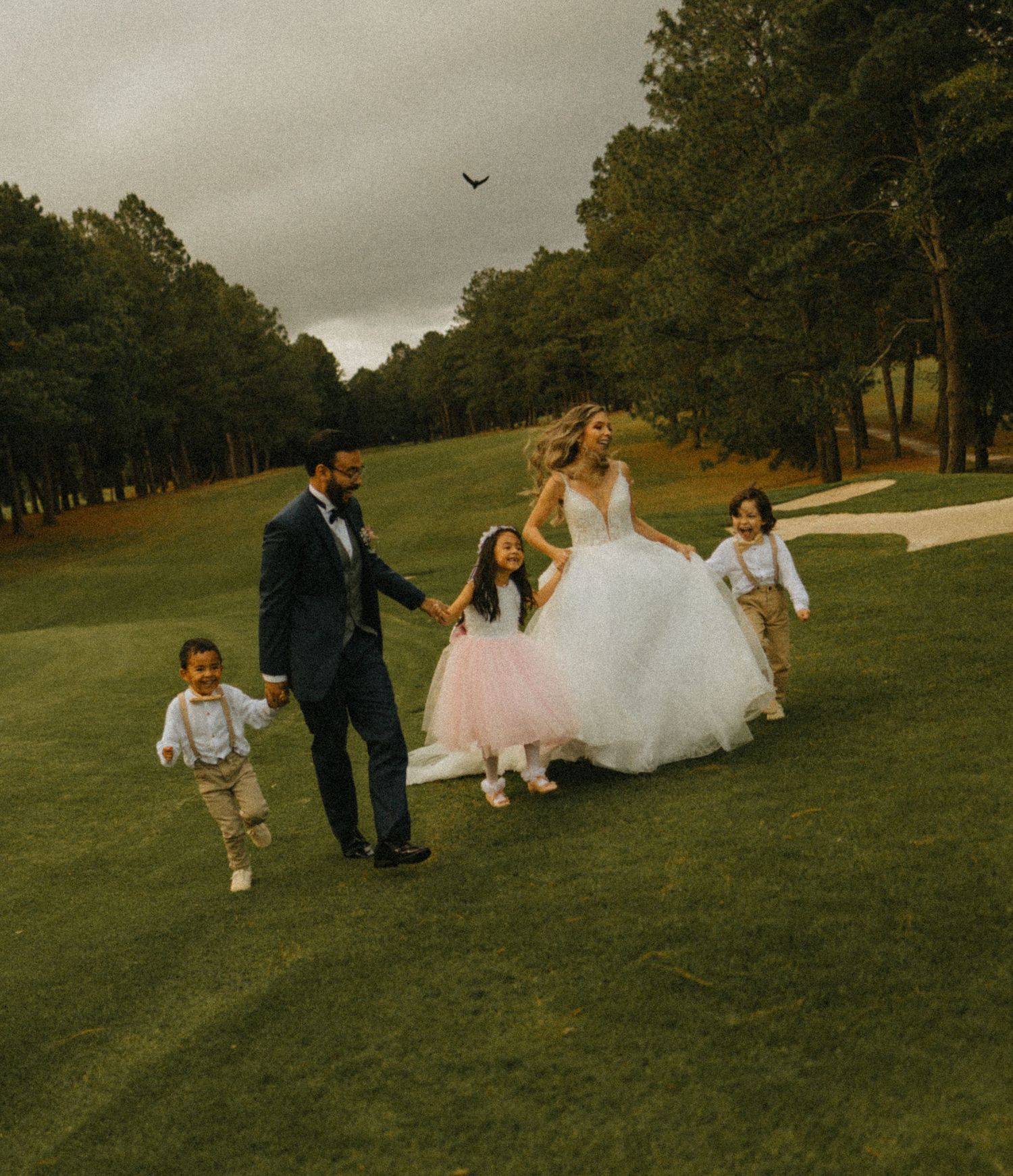 Bride and groom walk hand-in-hand with three children on a lush golf course, celebrating their wedding day outdoors.