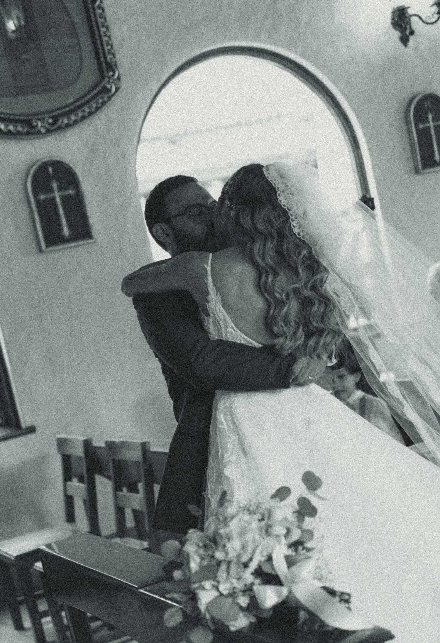 Black and white photo of a bride and groom sharing a kiss inside a church during their wedding ceremony.