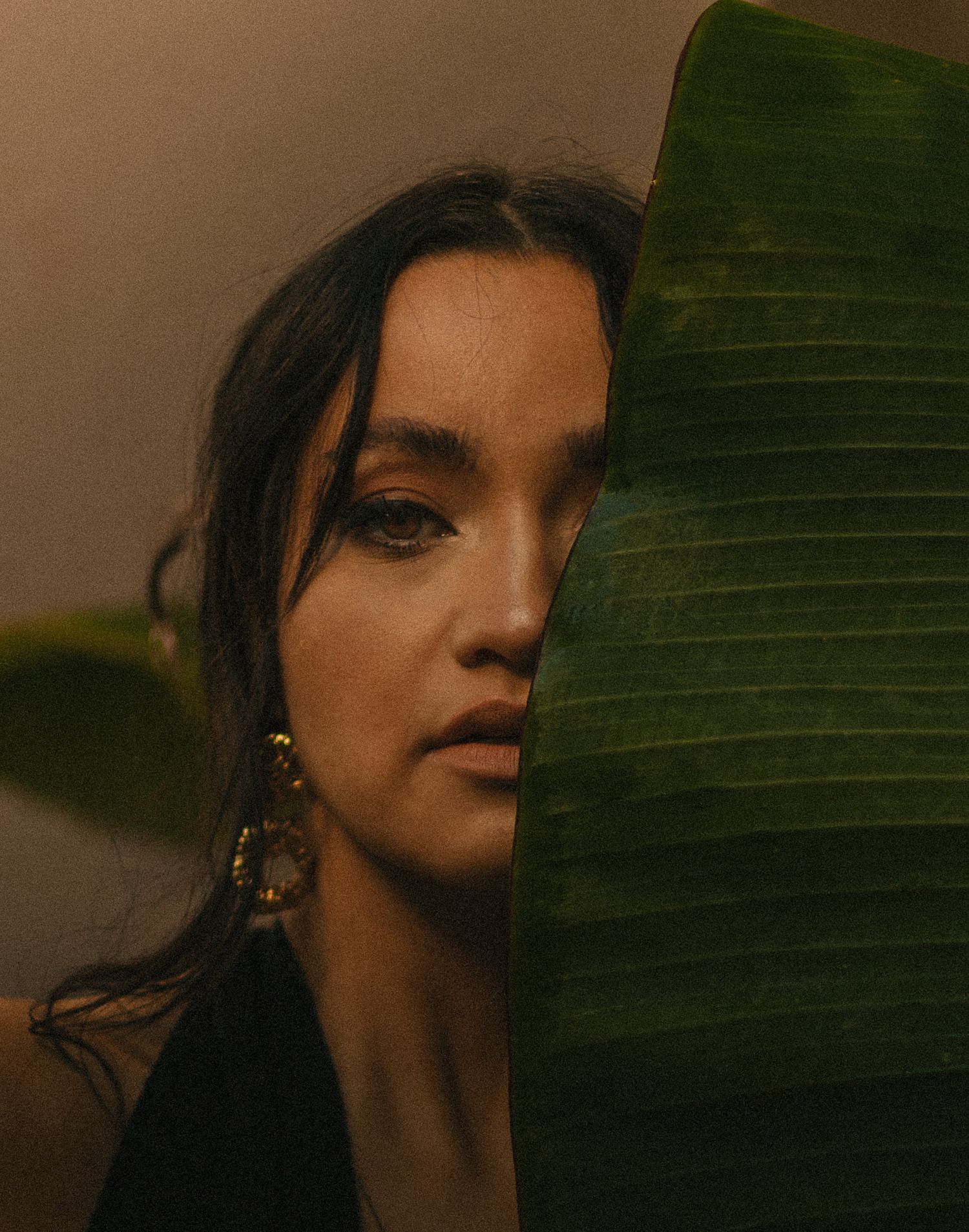 Young woman with dark hair and gold hoop earring peers from behind a large tropical leaf in moody lighting.