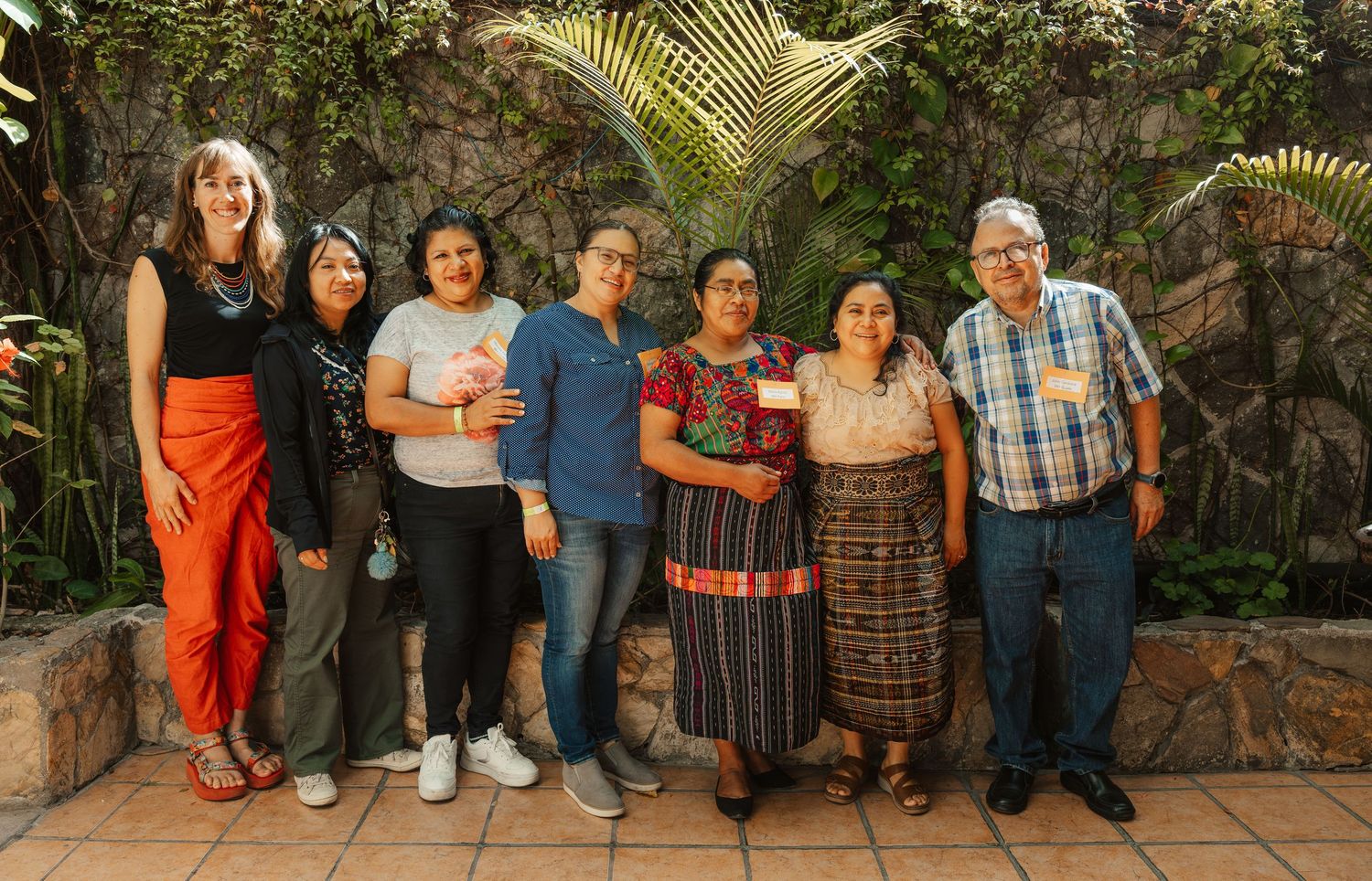 Group of seven people smiling together outdoors in a lush garden setting, some wearing traditional Guatemalan attire.