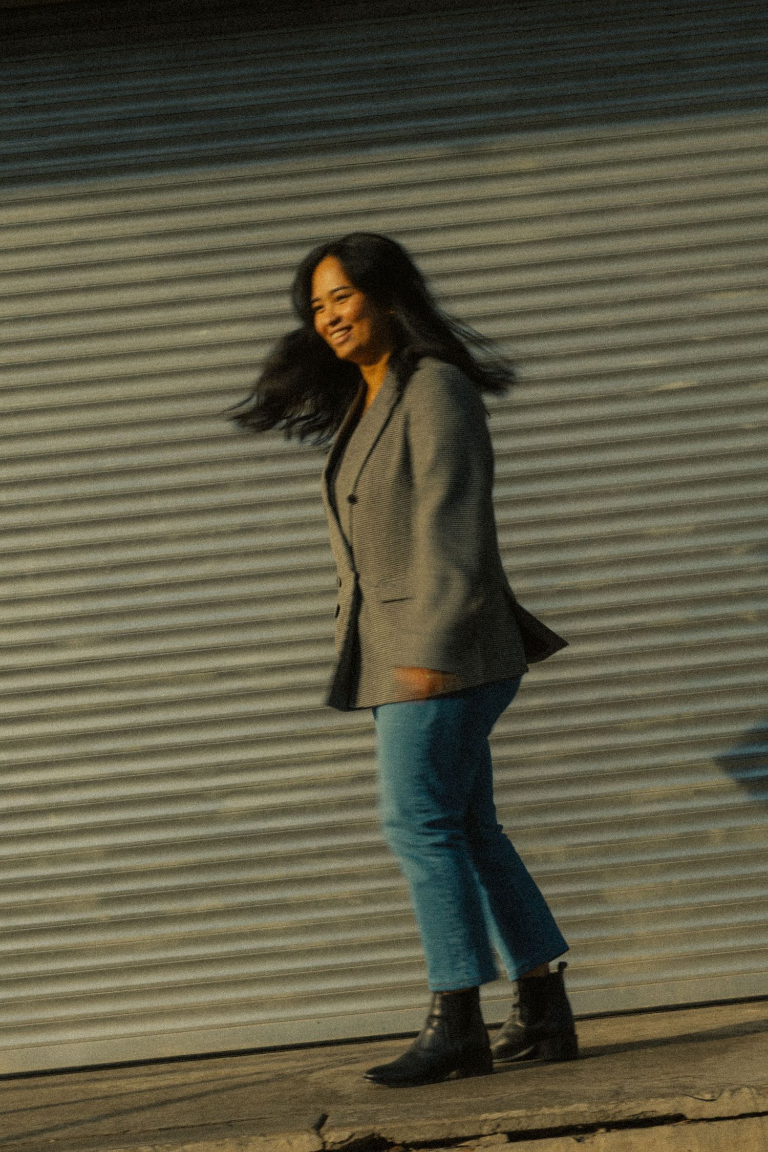 A smiling woman with flowing dark hair poses in a gray blazer, blue jeans, and black ankle boots against a metal wall.