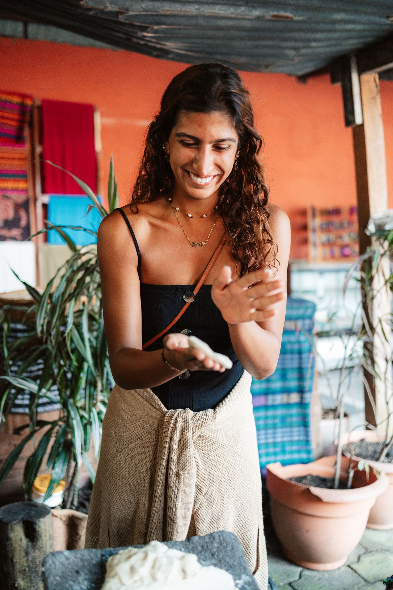 A smiling young woman with curly hair holding masa dough in a colorful Latin American market setting.