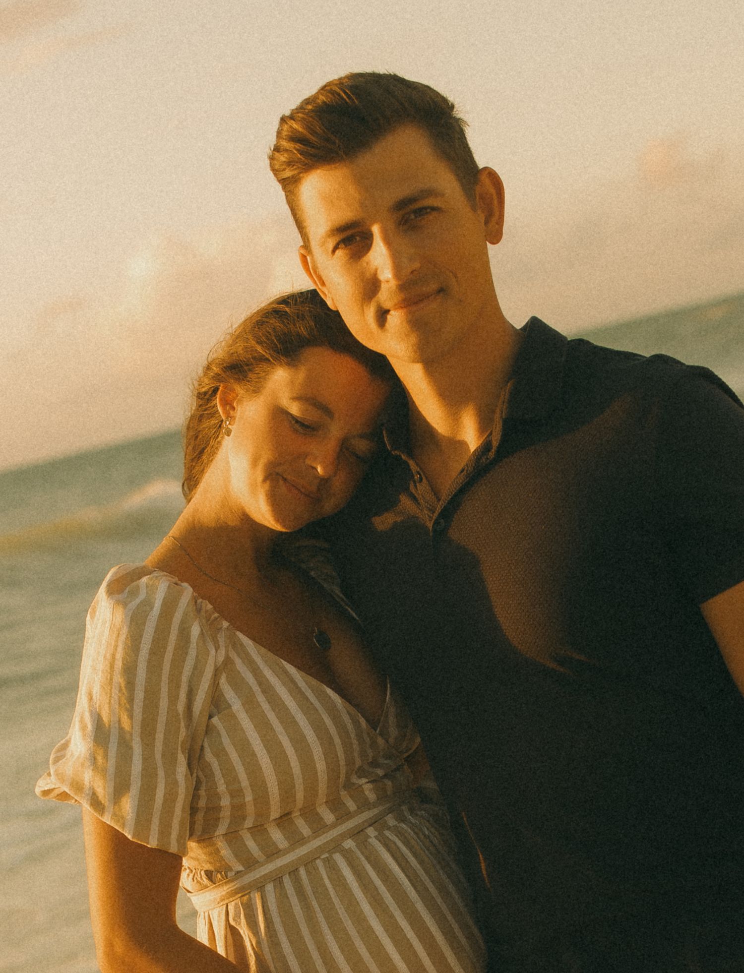 Couple embracing on a beach at sunset, woman in striped dress leaning on mans shoulder, golden hour light.