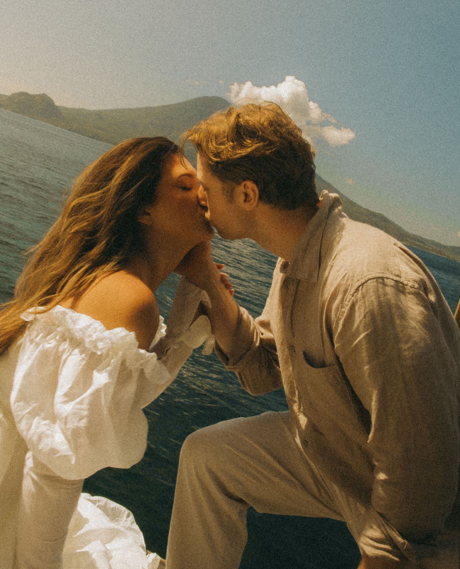 Couple sharing a romantic kiss on a boat with mountain lake scenery in the background.
