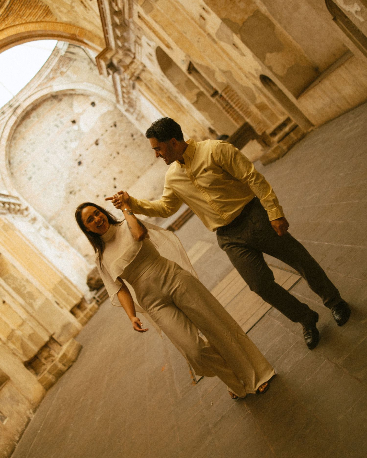 A couple dances joyfully in a historic stone archway, the woman in a white outfit and man in a yellow shirt.