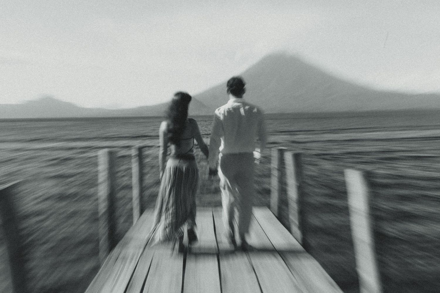 Black and white photo of a couple holding hands on a wooden dock overlooking a volcanic mountain and calm lake.