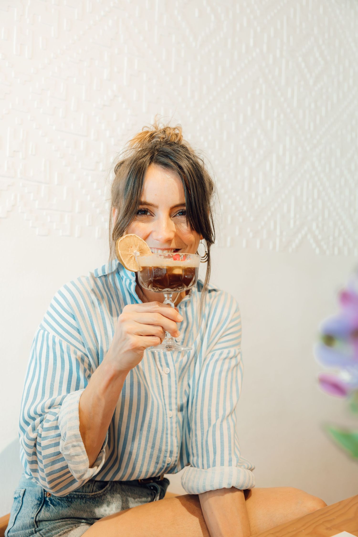 Smiling woman in a blue striped shirt and hair bun holds a cocktail garnished with a dried orange slice to her face.