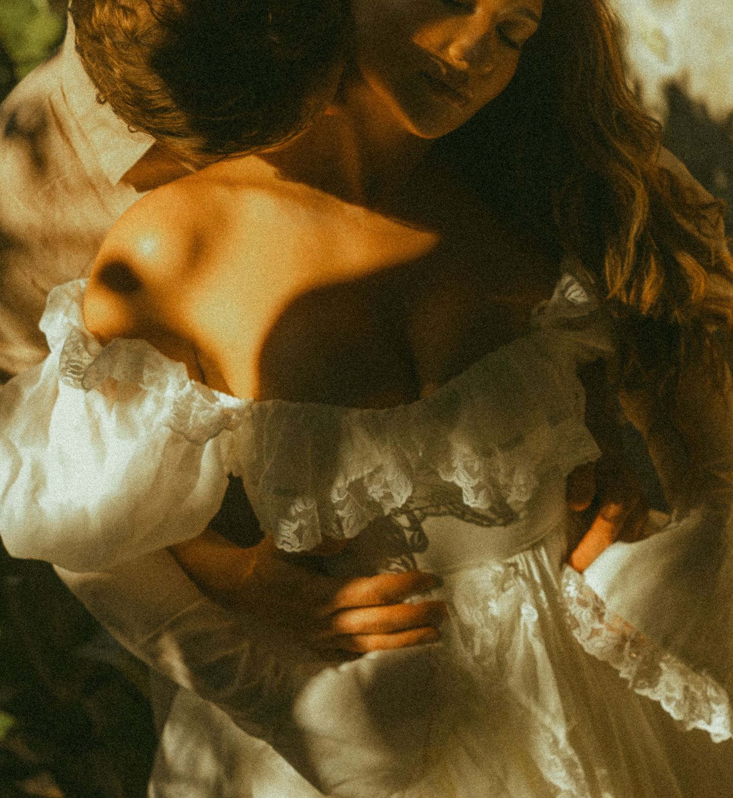 Bride in off-shoulder white lace wedding dress embraced by groom in warm sunlight.