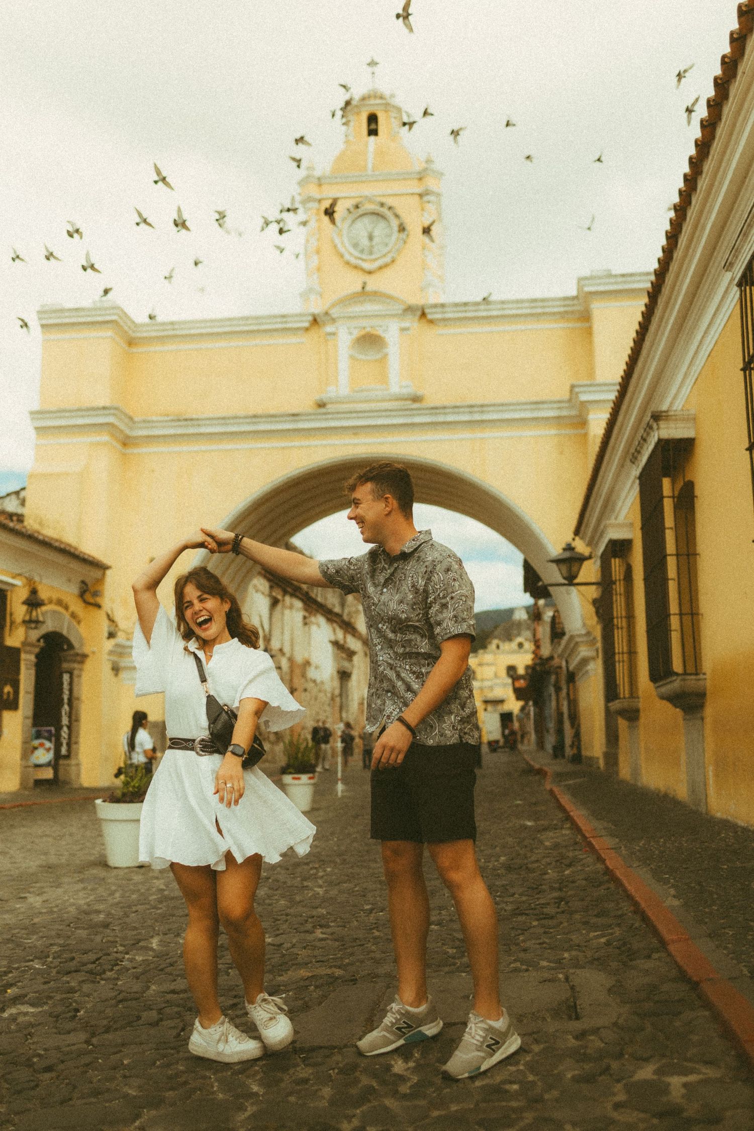 Couple dancing on cobblestone street in Antigua Guatemala with iconic yellow Santa Catalina arch and clock tower behind them.