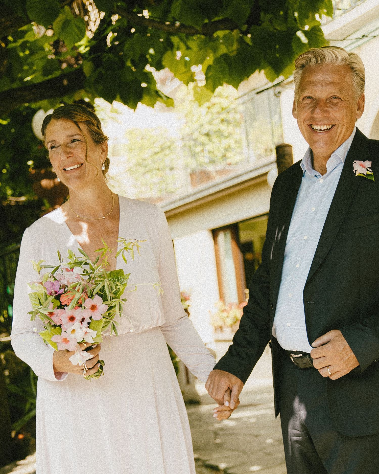 Smiling bride in white dress holding pink bouquet walks hand in hand with groom in dark suit outdoors.