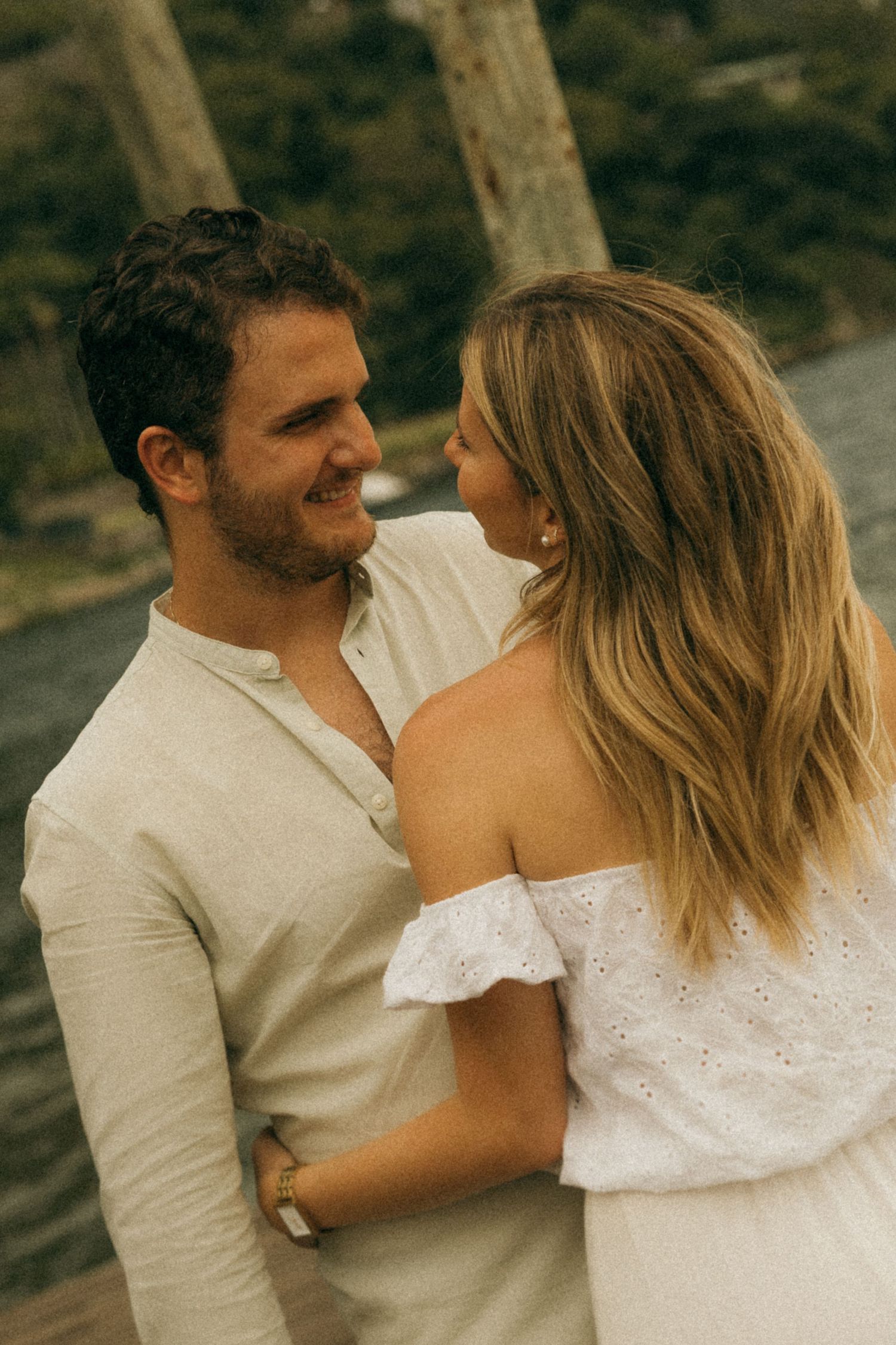 A couple sharing a romantic moment outdoors, man in cream shirt smiling at blonde woman in white off-shoulder dress.