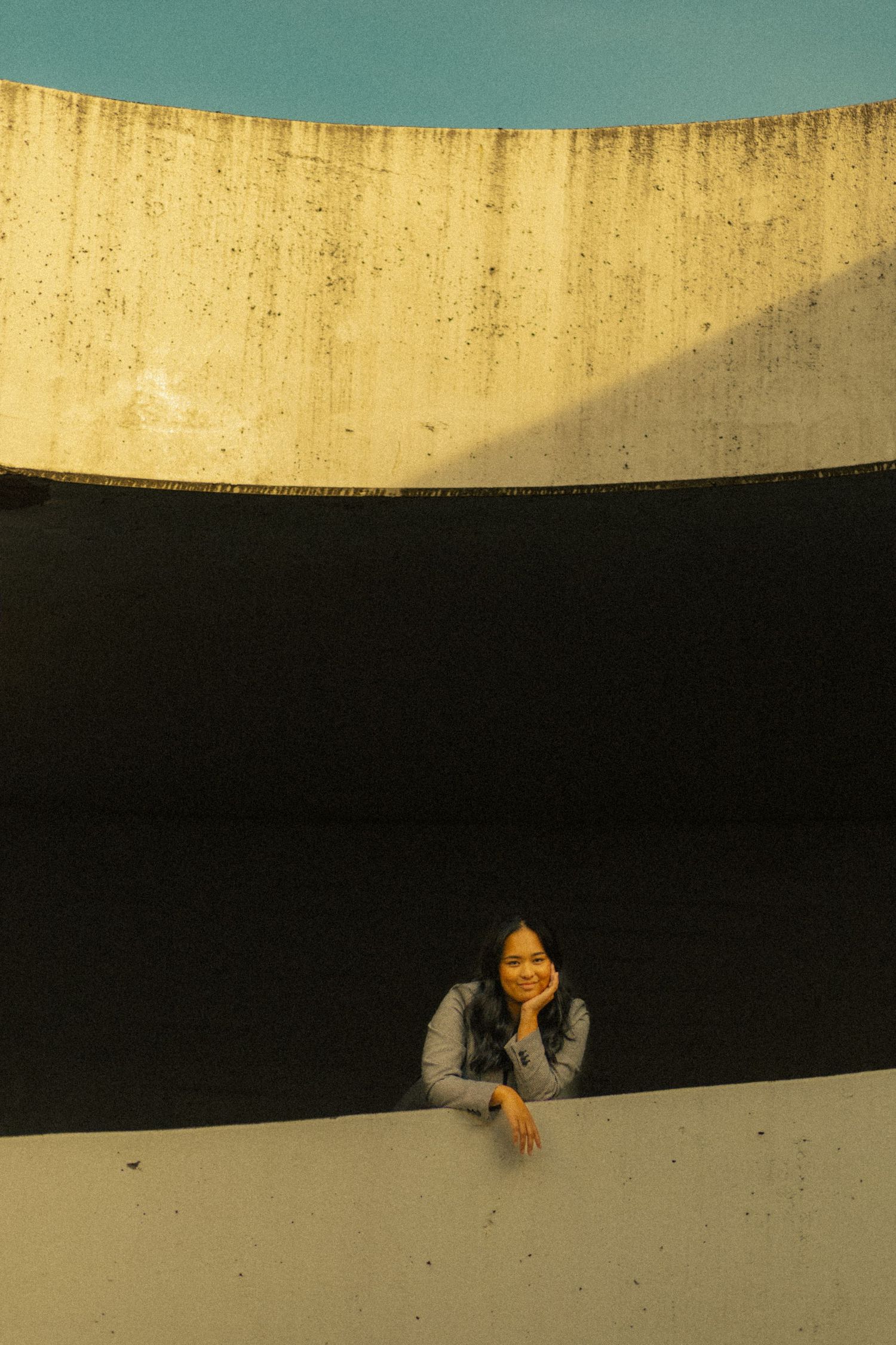 A smiling young woman leans on a ledge against a dramatic curved concrete wall with contrasting light and shadow.