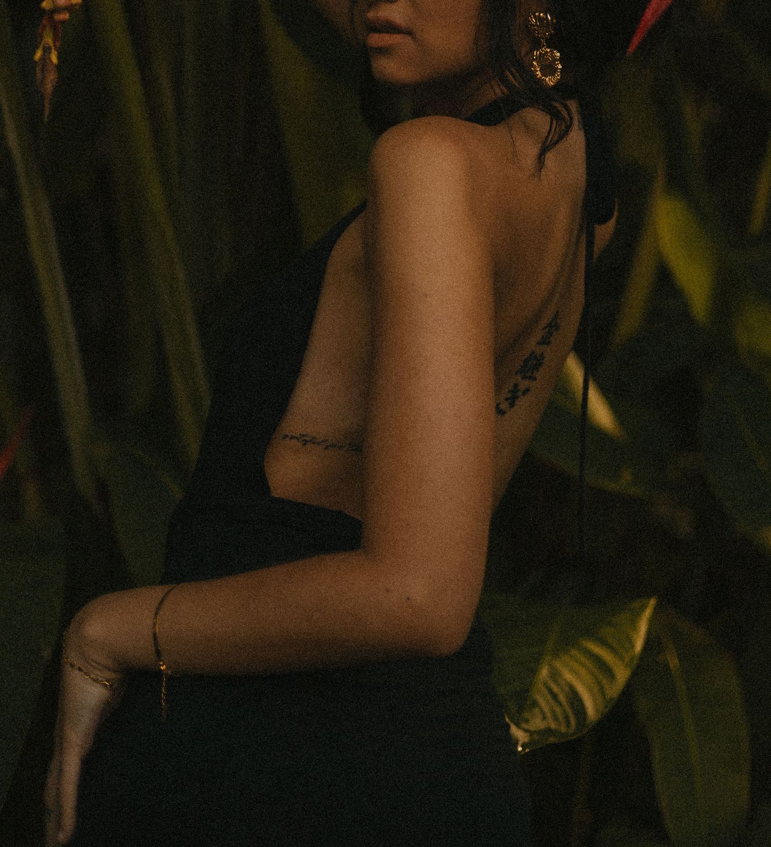 Woman in black backless dress with gold jewelry and tattoo, surrounded by tropical green foliage.