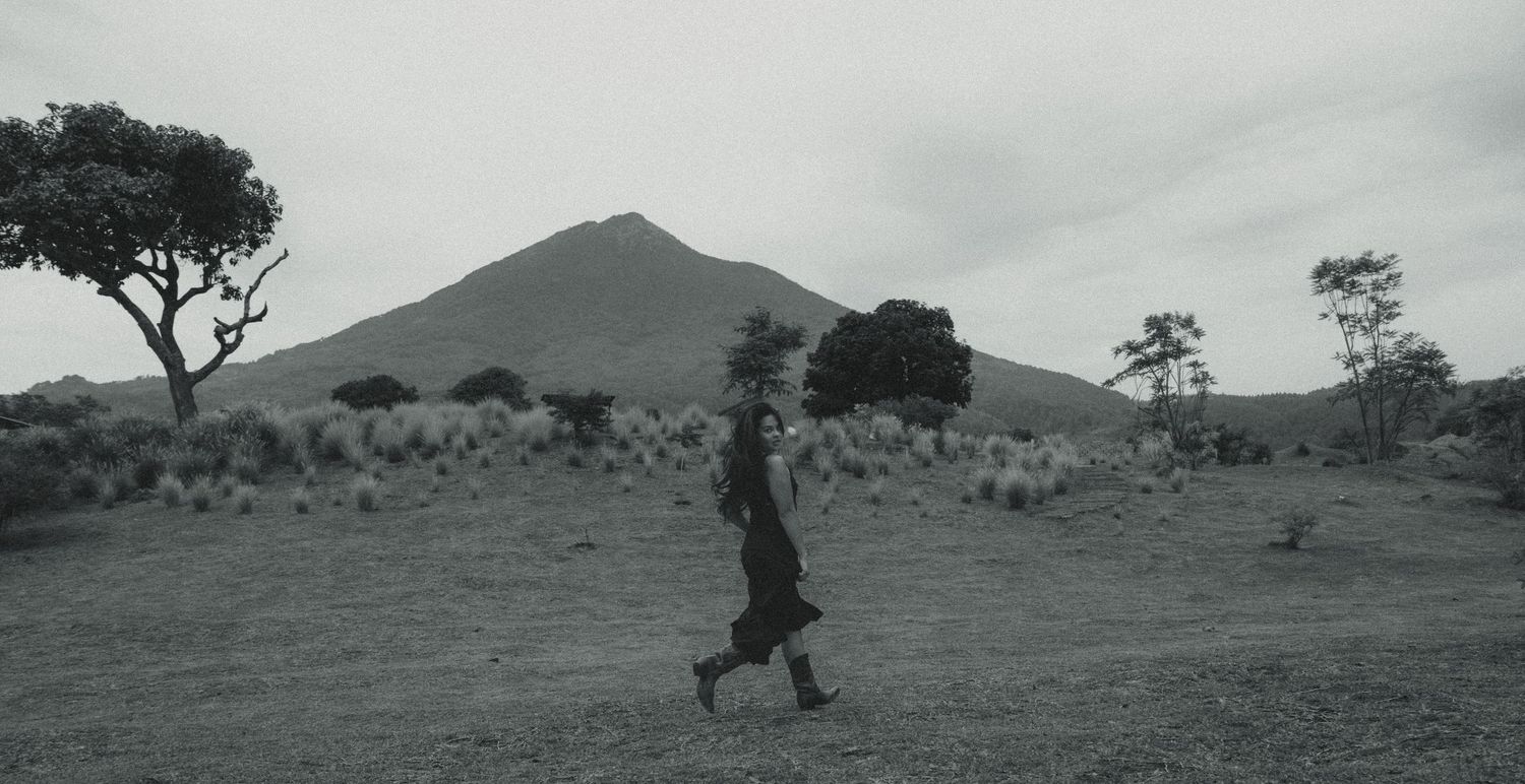 Black and white photo of a person walking across an open field with a volcanic mountain in the background.