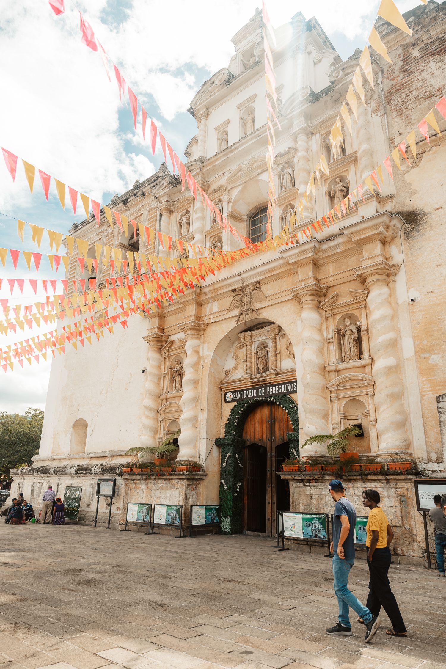 Two people walk past a historic baroque church with an ornate facade, decorated with colorful papel picado banners in a sunny plaza.