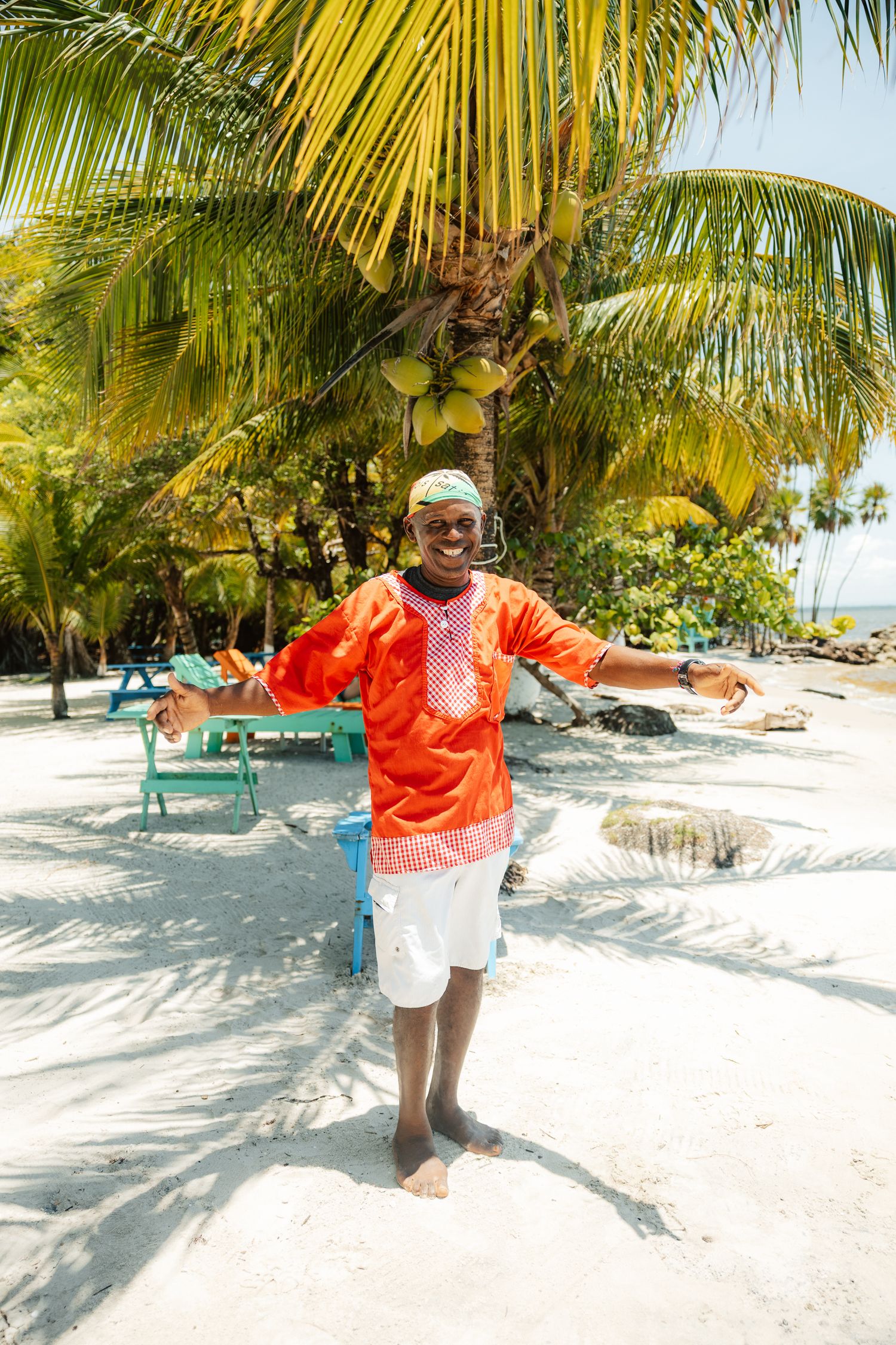 Smiling man in orange traditional shirt stands on white sand beach with palm trees in tropical paradise.