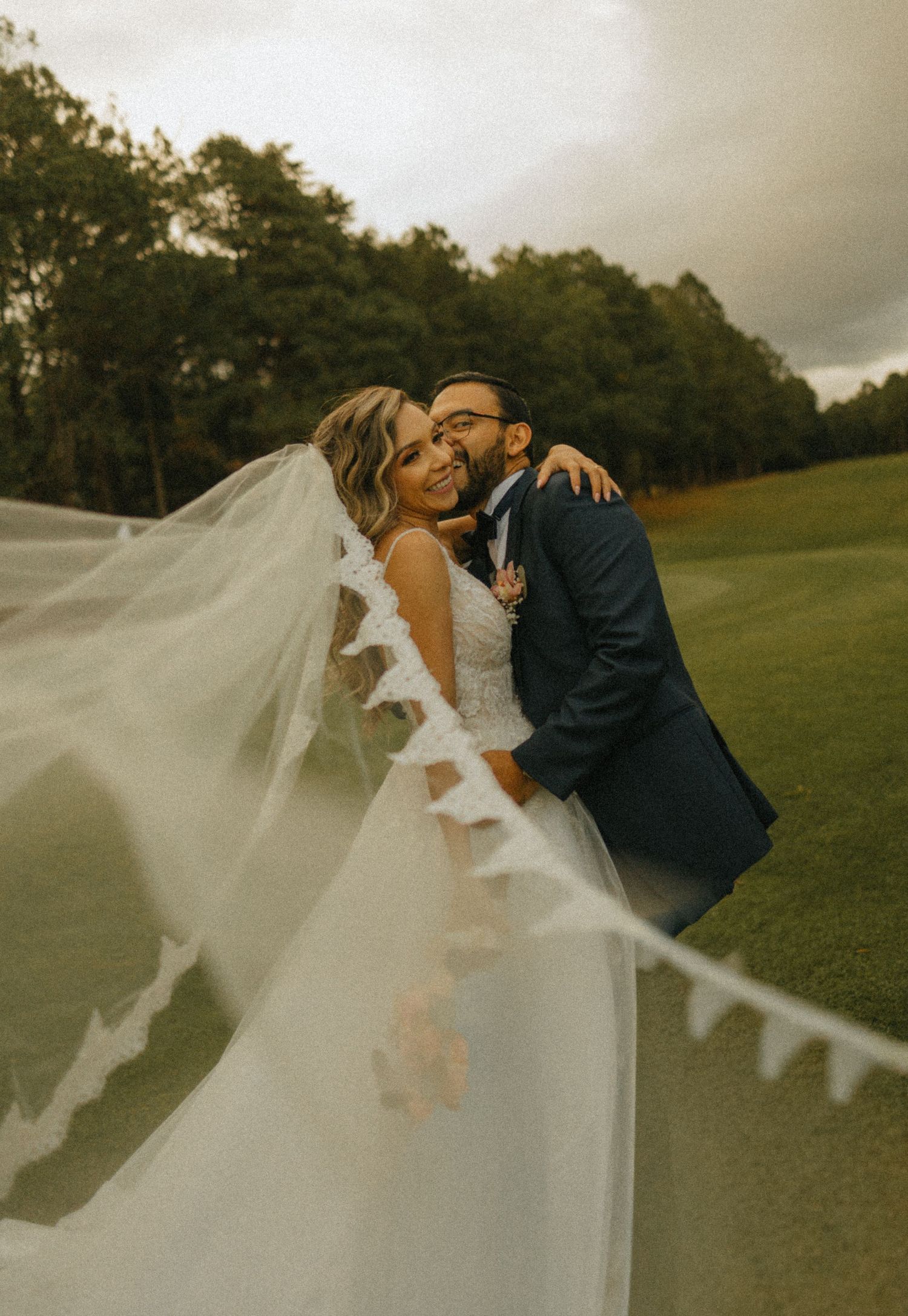 Bride and groom embracing on golf course, veil billowing in wind, wearing white gown and navy suit on cloudy day.