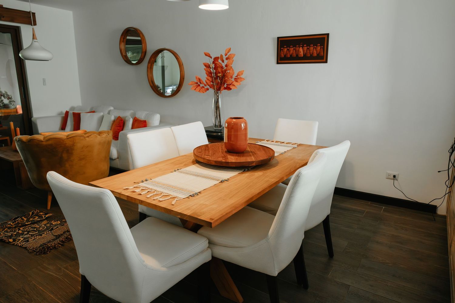 Elegant dining room with wooden table, white upholstered chairs, orange floral centerpiece, and round mirrors on gray wall.