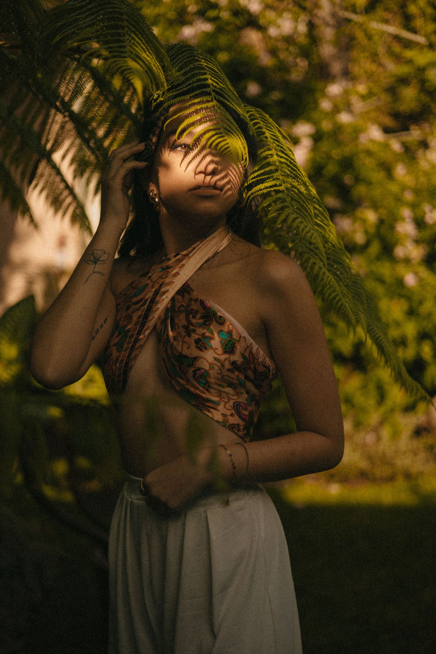 Woman in floral top and white pants poses with tropical fern leaves in a lush garden setting.