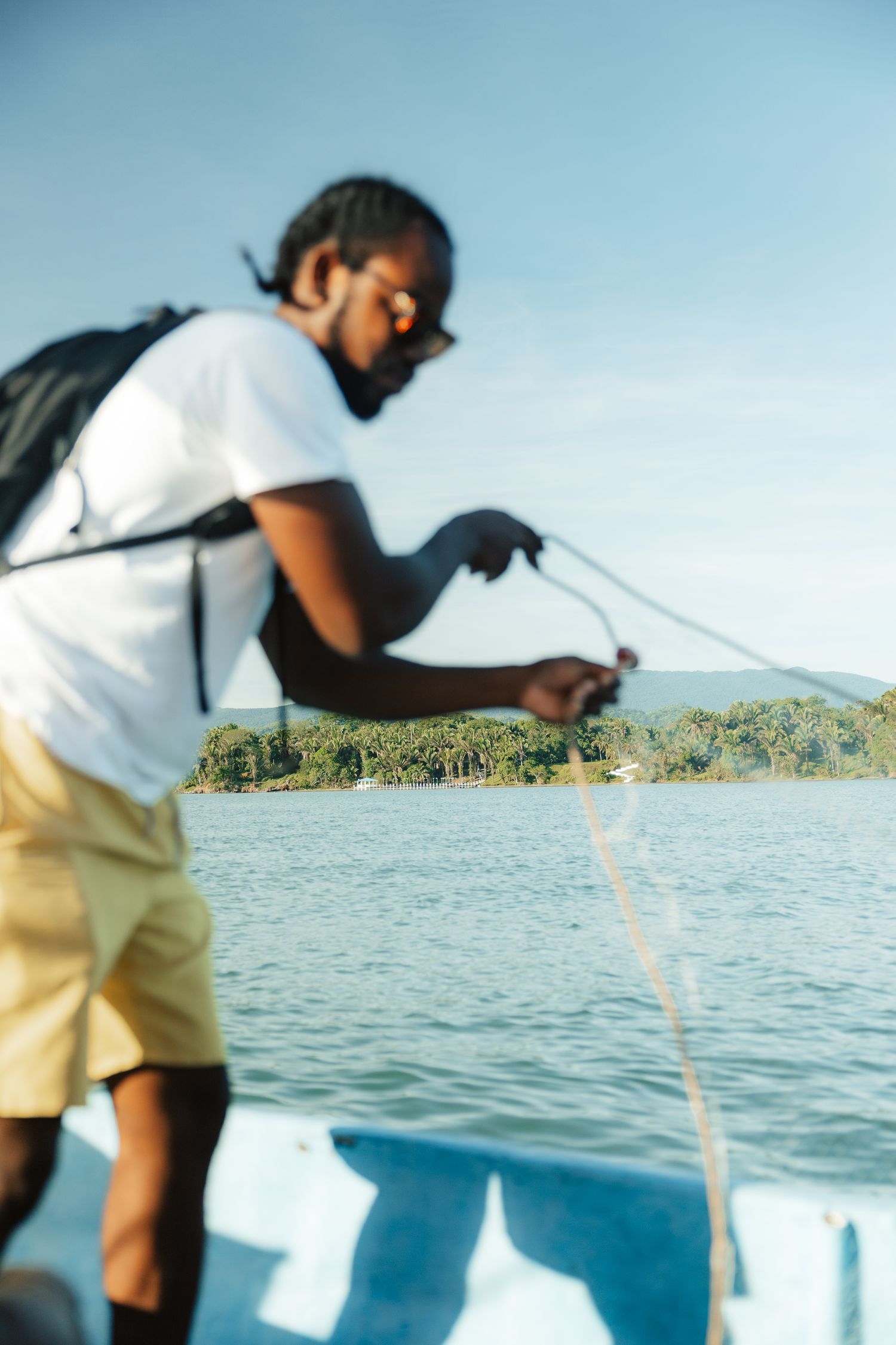 A Black man in a white shirt and yellow shorts handles a rope on a boat with tropical greenery and blue water in the background.