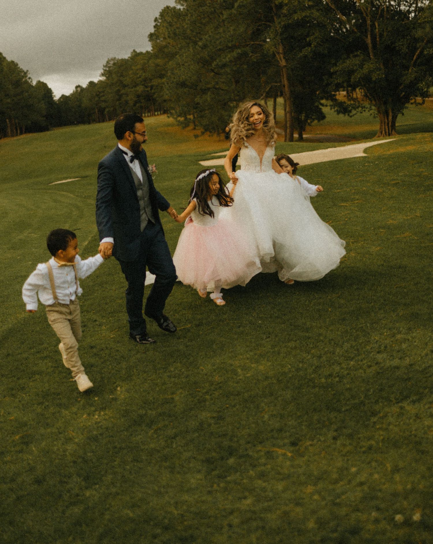 Bride and groom in wedding attire joyfully running on green grass with two children in formal wear.