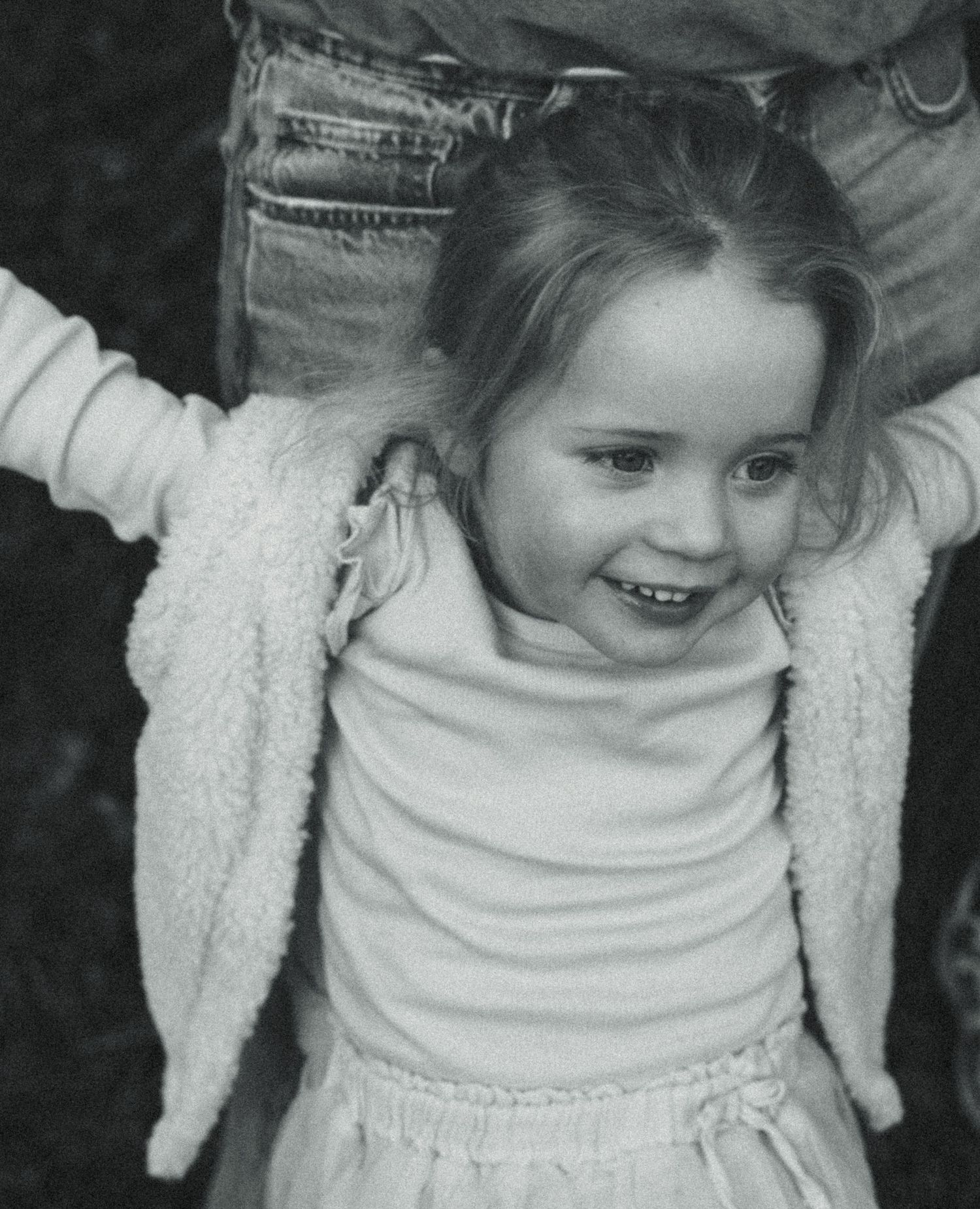 A smiling young girl in a white fleece jacket being held up by an adult outdoors in a black and white photo.