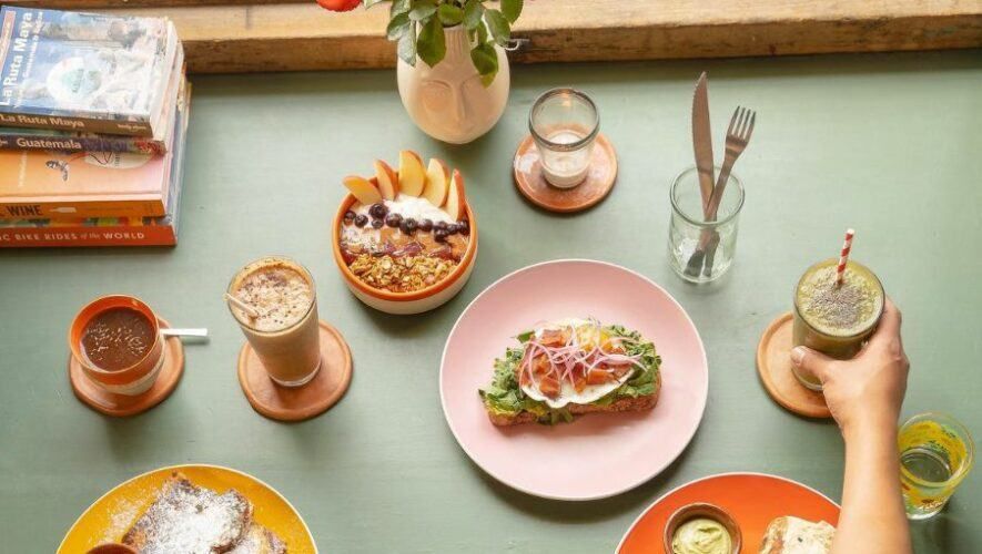 Overhead view of a colorful brunch spread with avocado toast, granola bowls, smoothies, and coffee on a green table.