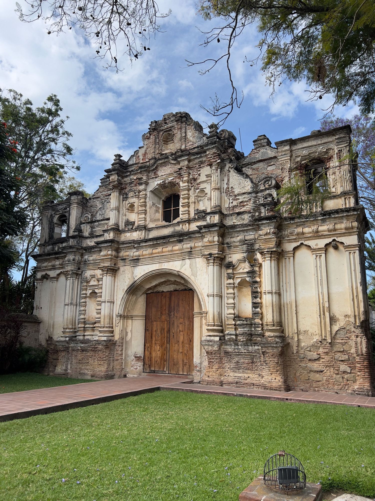 Historic baroque church ruins with ornate stone facade, wooden doors, and lush green lawn in Antigua, Guatemala.