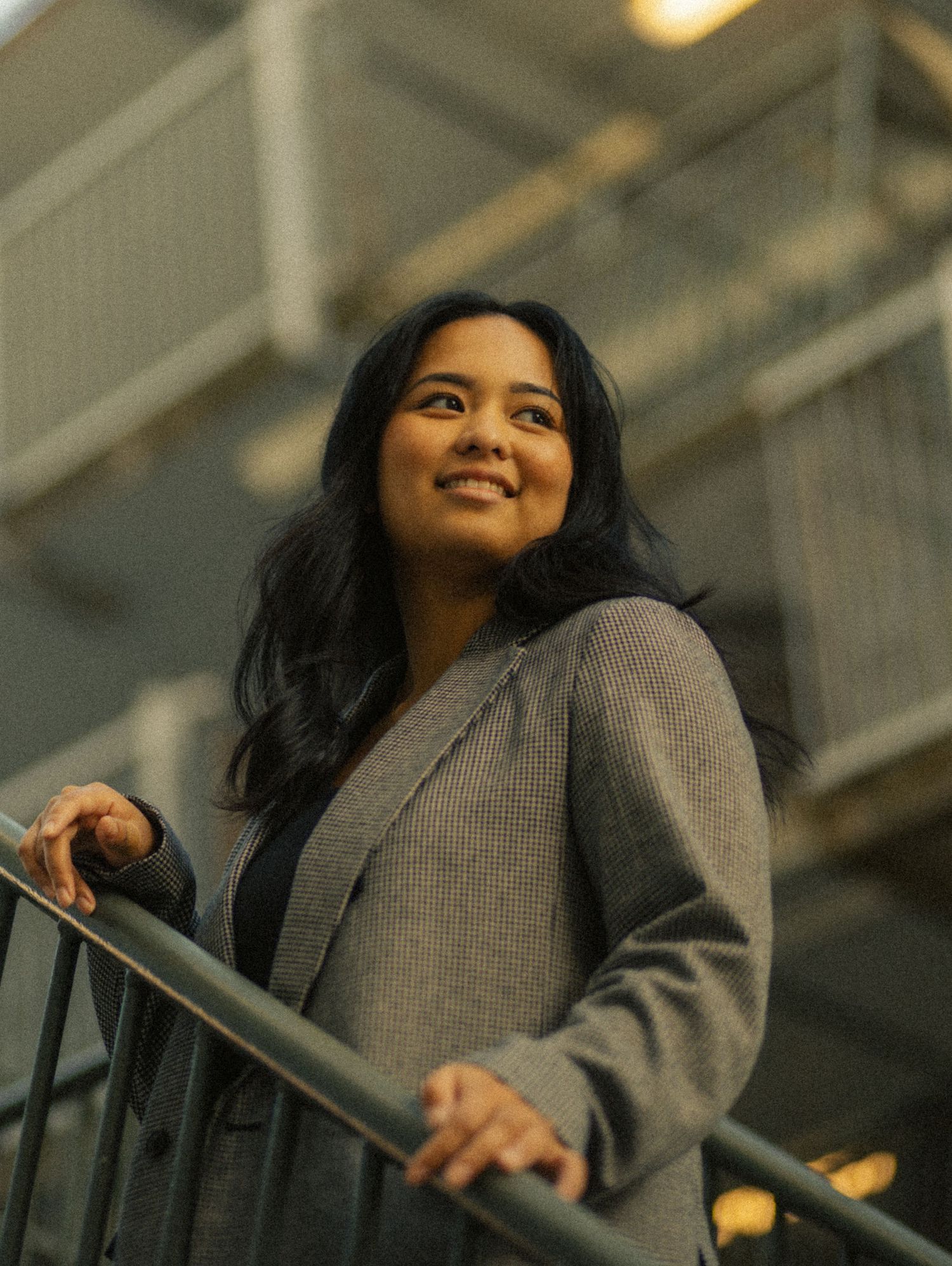 Young woman in gray blazer smiling confidently while leaning on a railing in a modern building interior.